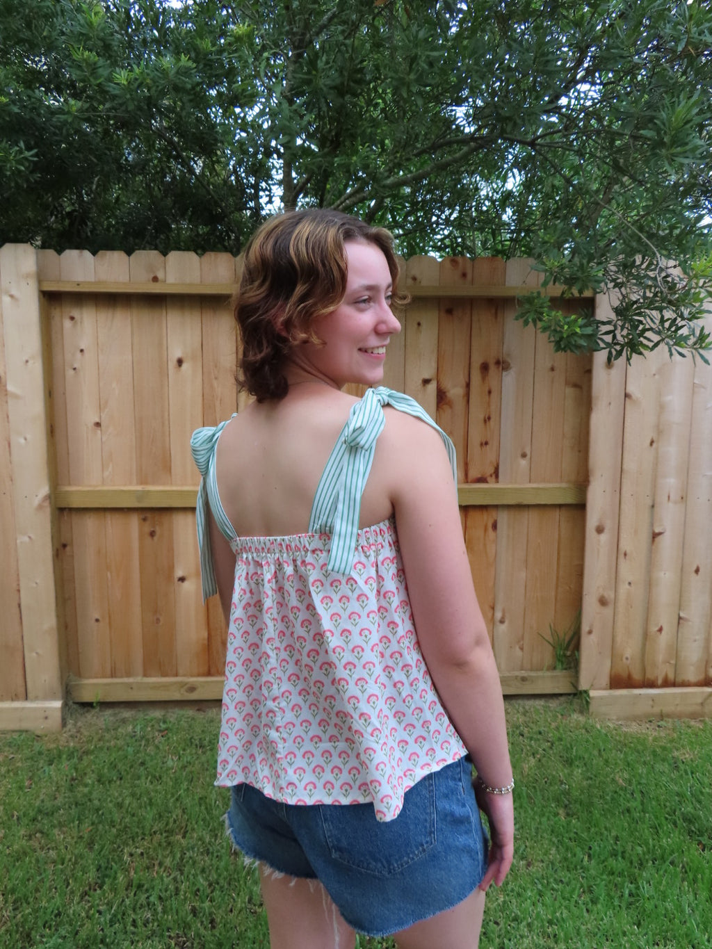 A woman wearing a sleeveless tie-strap top with a pink and white floral pattern and light green and white striped shoulder straps, paired with denim shorts, standing outdoors in front of a wooden fence and green trees.
