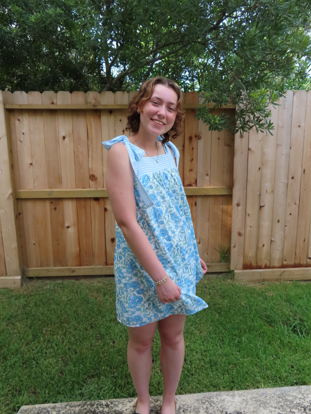 A young girl wearing a light blue and white floral print sleeveless dress, standing outdoors in front of a wooden fence and green trees.
