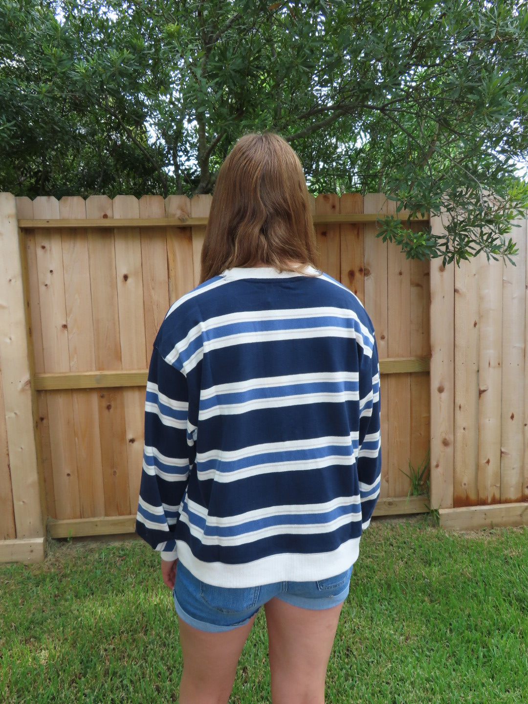 A person viewed from behind wearing a navy blue and white striped t-shirt and denim shorts, standing outdoors in front of a wooden fence with green grass and trees.