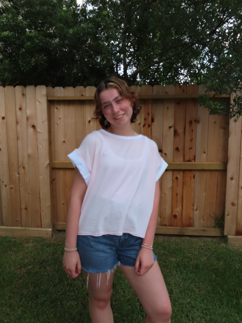 A young woman smiling and posing outdoors in front of a wooden fence, wearing a pink short-sleeve t-shirt and denim shorts.