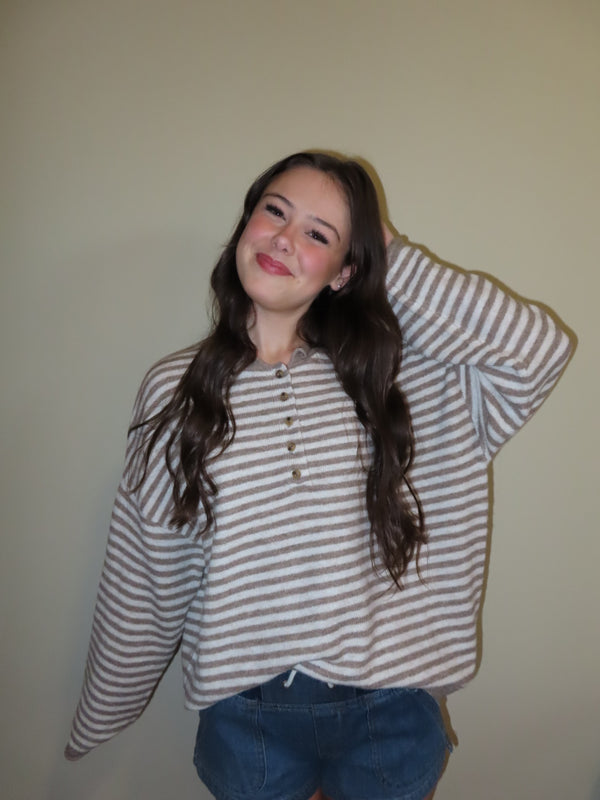 A young woman with long wavy brown hair wearing a beige and white horizontally striped long-sleeve henley shirt and blue jean shorts, posing against a plain beige wall.