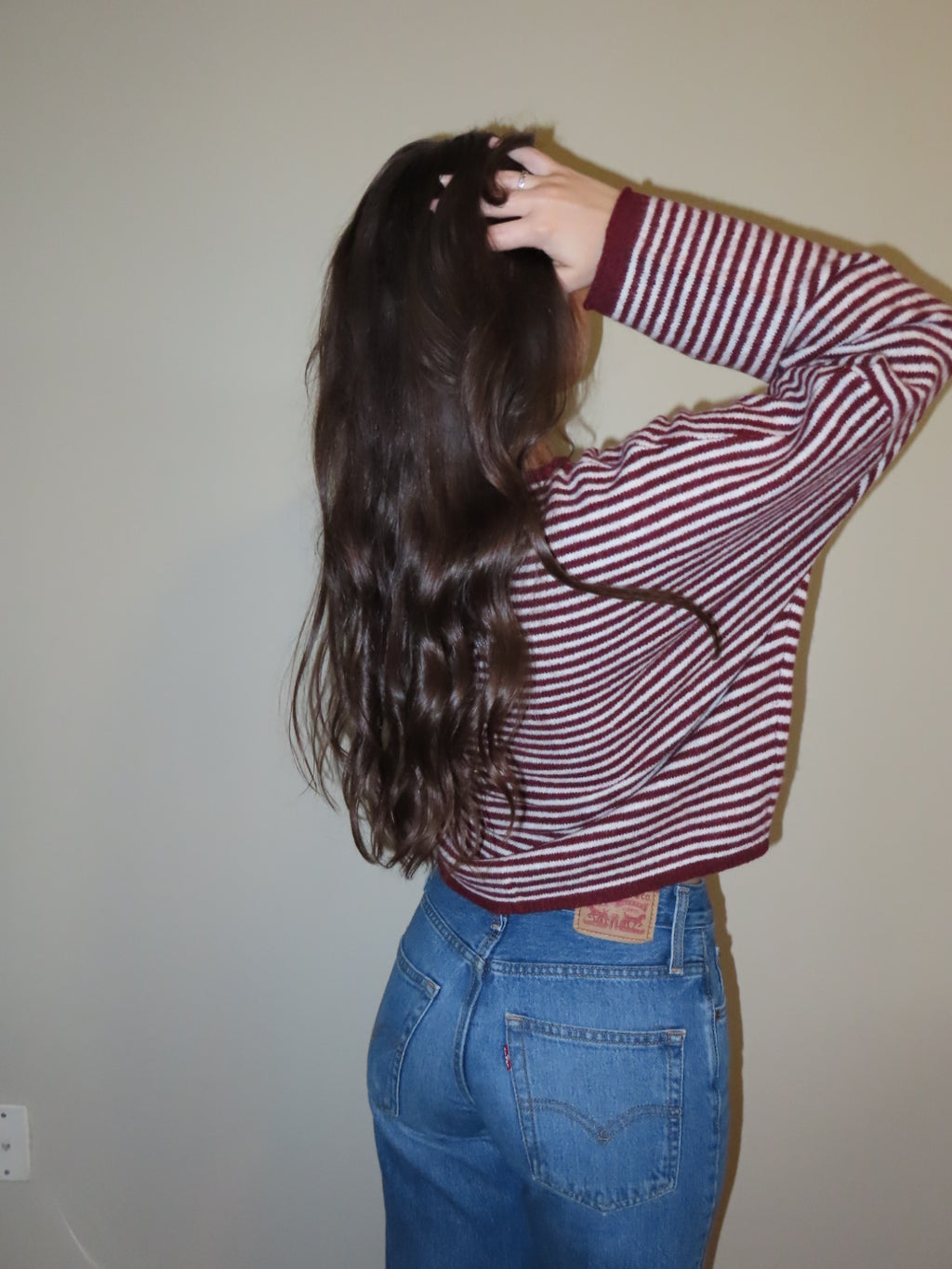 A person with long, wavy dark brown hair is shown from the back, wearing a red and white horizontally striped long-sleeve crop top and blue jeans, standing against a plain beige wall.