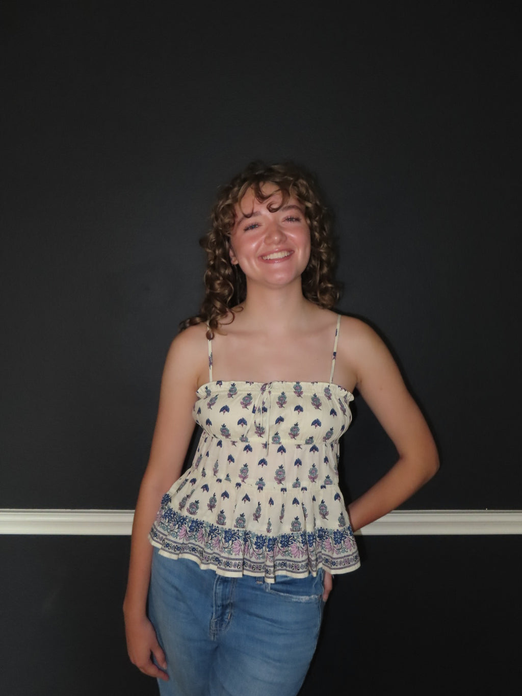 A smiling young woman with curly hair stands against a dark wall wearing a cream-colored spaghetti-strap top with a blue and purple floral pattern, paired with blue jeans.