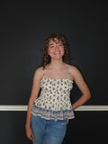 A smiling young woman with curly hair stands against a dark wall wearing a cream-colored spaghetti-strap top with a blue and purple floral pattern, paired with blue jeans.