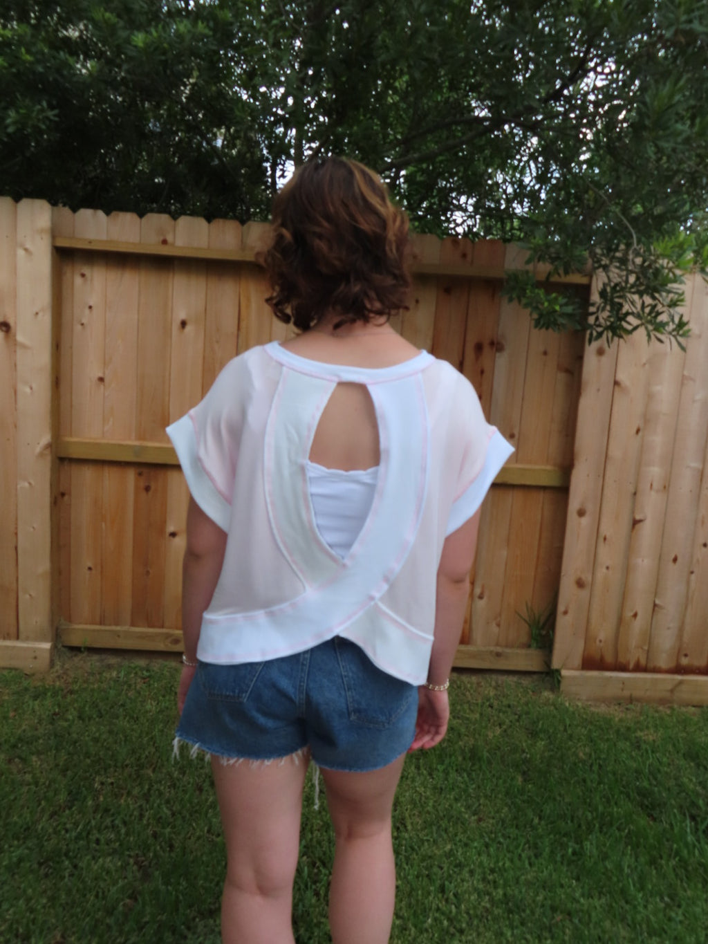 A person standing outdoors in front of a wooden fence and grass, wearing a pink short sleeve top with a large open cutout in the back and denim shorts.