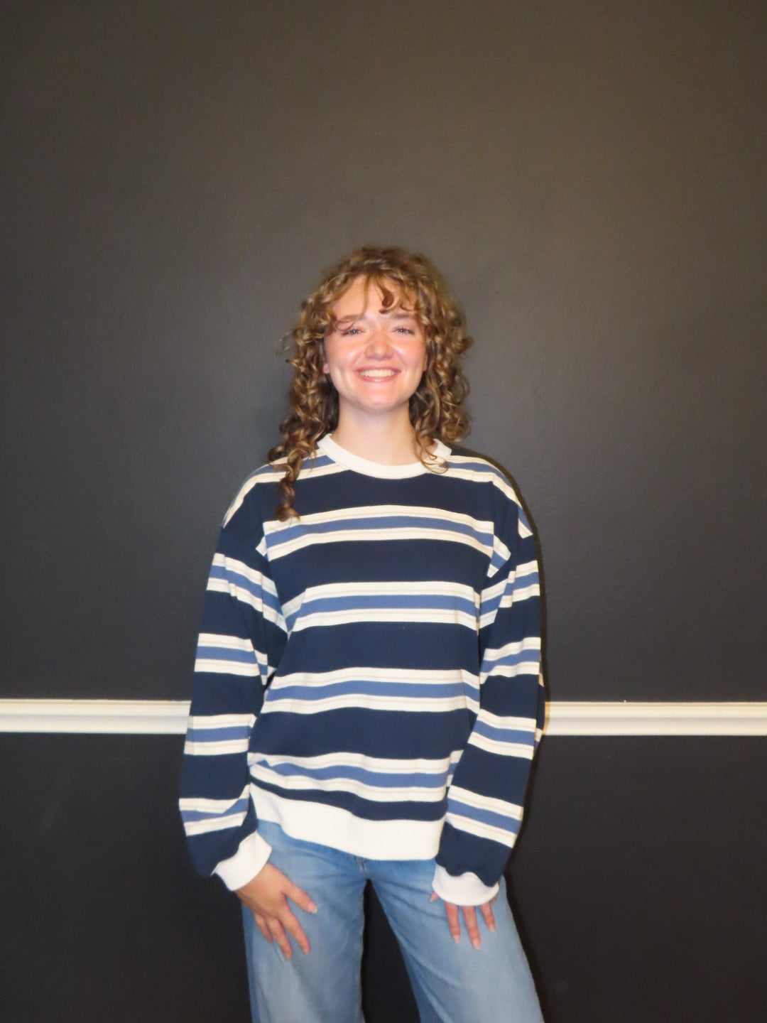 A smiling young woman with curly hair stands against a dark gray wall wearing a navy blue and white horizontally striped long-sleeve sweater and light blue jeans.