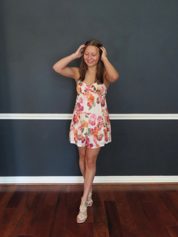 A woman modeling a sleeveless white mini dress with a colorful floral print, standing against a dark gray wall with a white chair rail and dark wood floors.