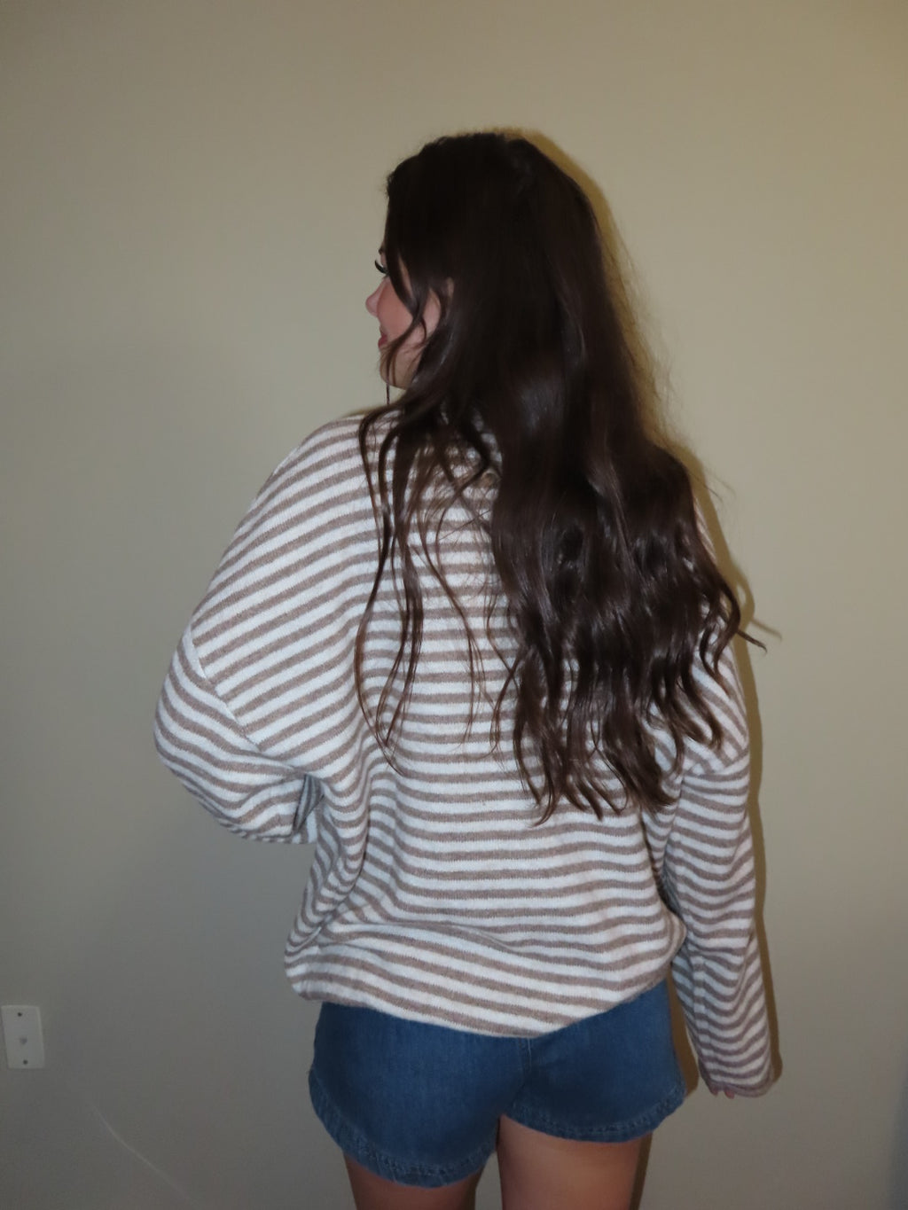 A person with long dark hair, viewed from behind, wearing a beige and white striped long-sleeve shirt and blue denim shorts against a plain beige wall.
