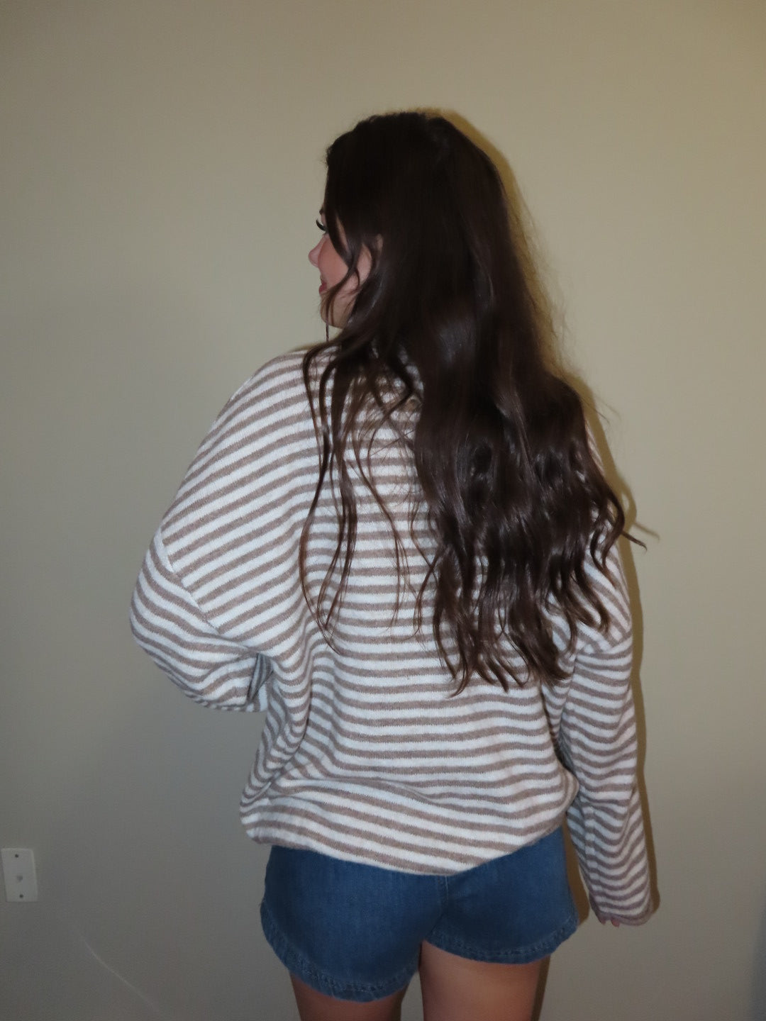 A person with long dark hair, viewed from behind, wearing a beige and white striped long-sleeve shirt and blue denim shorts against a plain beige wall.