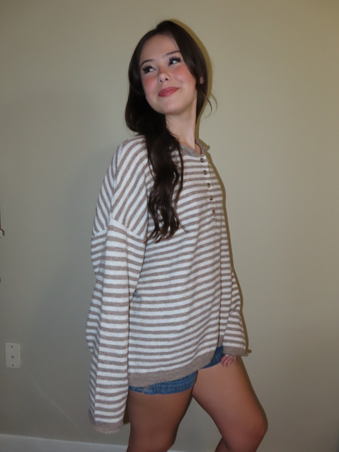 A woman wearing an oversized beige and white horizontally striped henley sweatshirt with denim shorts, standing against a plain beige wall.