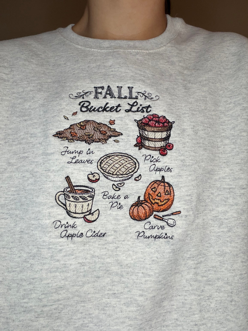 A close-up of a person wearing a light heather gray crewneck sweatshirt with a 'Fall Bucket List' graphic printed on the chest. There is embroidered leaves, apples, a pie, apple cider, and pumpkin carving.