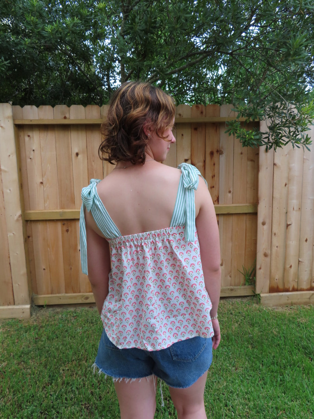 A woman wearing a sleeveless tie-strap top with a pink and white floral pattern and light green and white striped shoulder straps, paired with denim shorts, standing outdoors in front of a wooden fence and green trees.