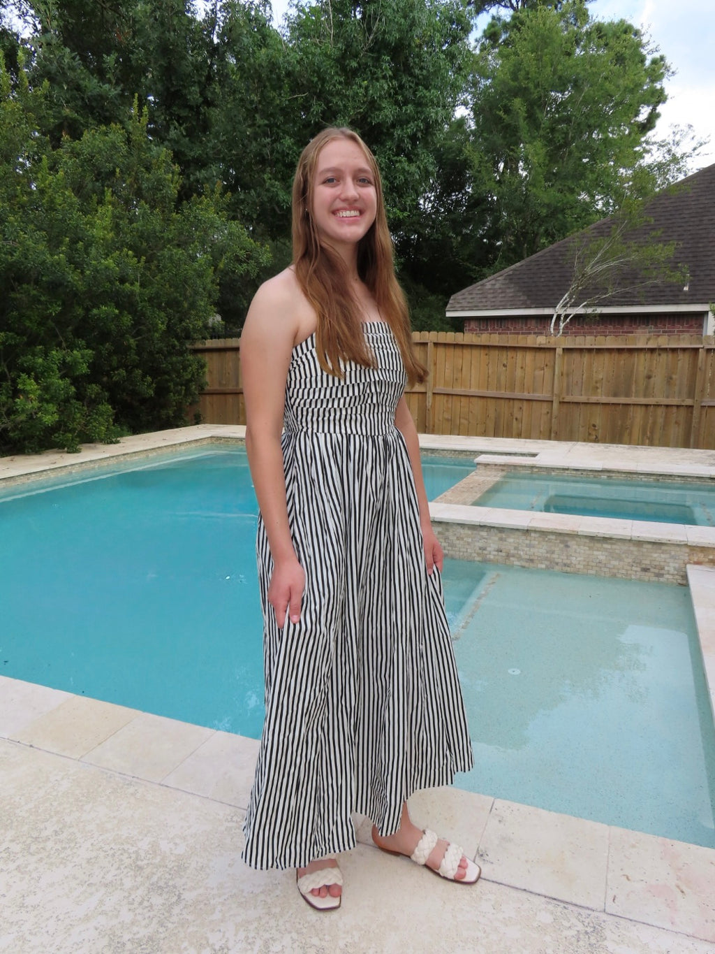 A woman wearing a black and white vertically striped strapless maxi dress stands next to a swimming pool in a backyard with trees and a wooden fence.