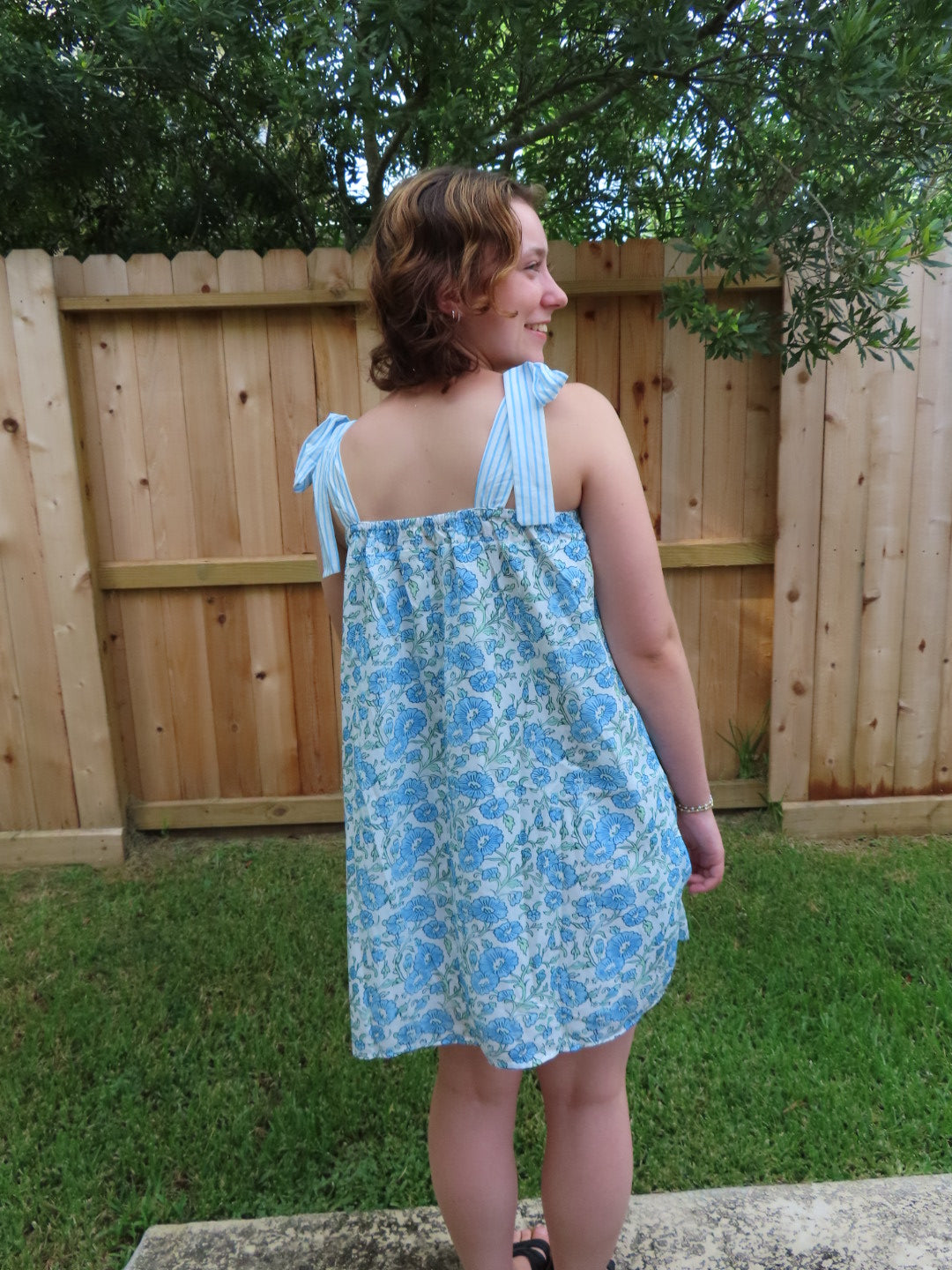 A woman pictured from behind wearing a light blue and white floral print sleeveless dress, standing outdoors in front of a wooden fence and green grass.