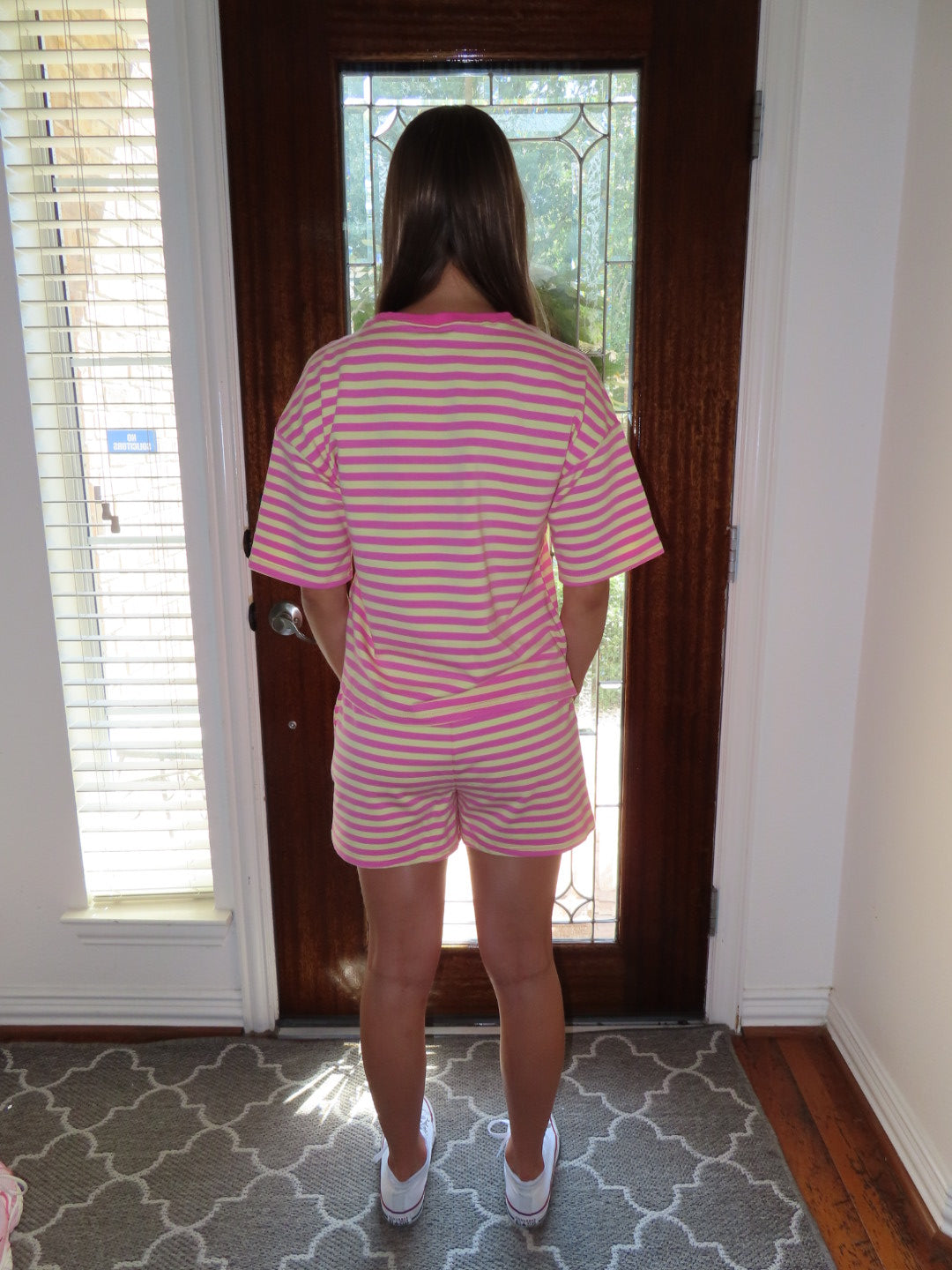A person viewed from behind wearing a pink and yellow striped short-sleeve top and matching shorts, standing in front of a dark wood front door with a window.