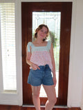 A young woman stands in front of a dark wood door wearing a light green and pink patterned sleeveless top with ruffled straps and blue denim shorts.