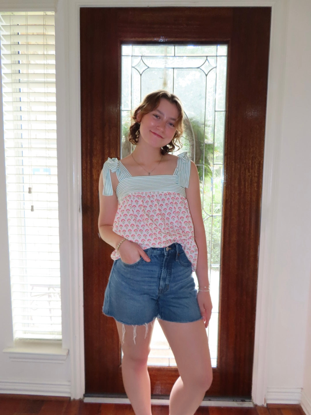 A young woman stands in front of a dark wood door wearing a light green and pink patterned sleeveless top with ruffled straps and blue denim shorts.