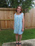 A young girl wearing a light blue and white floral patterned sleeveless dress, standing outdoors in front of a wooden fence and grass.