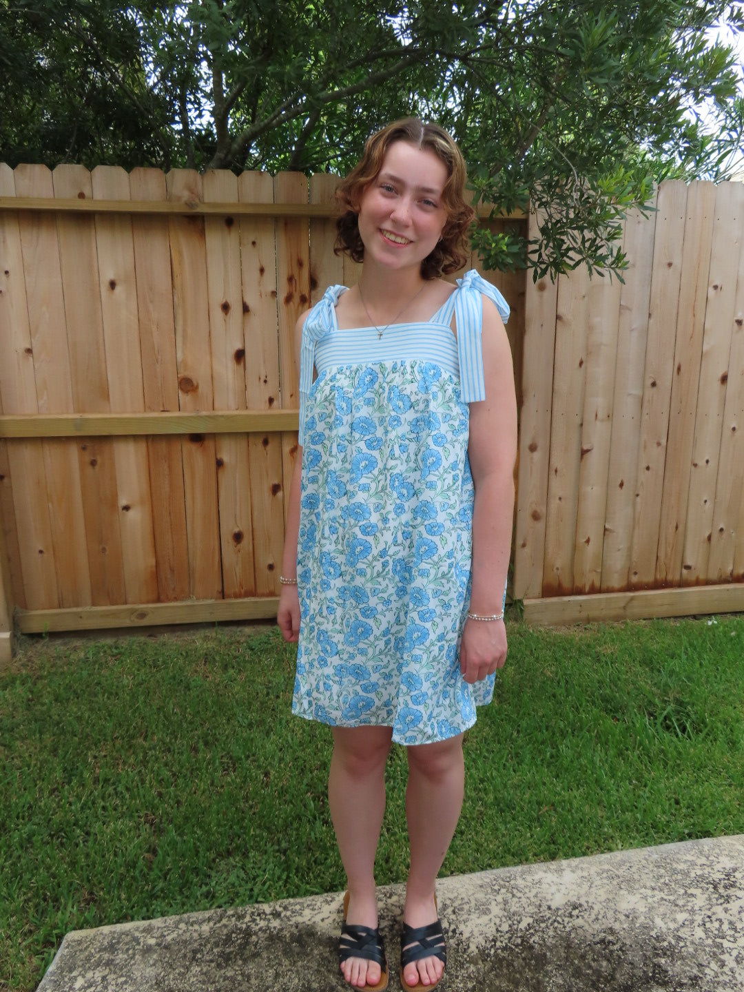 A young girl wearing a light blue and white floral patterned sleeveless dress, standing outdoors in front of a wooden fence and grass.