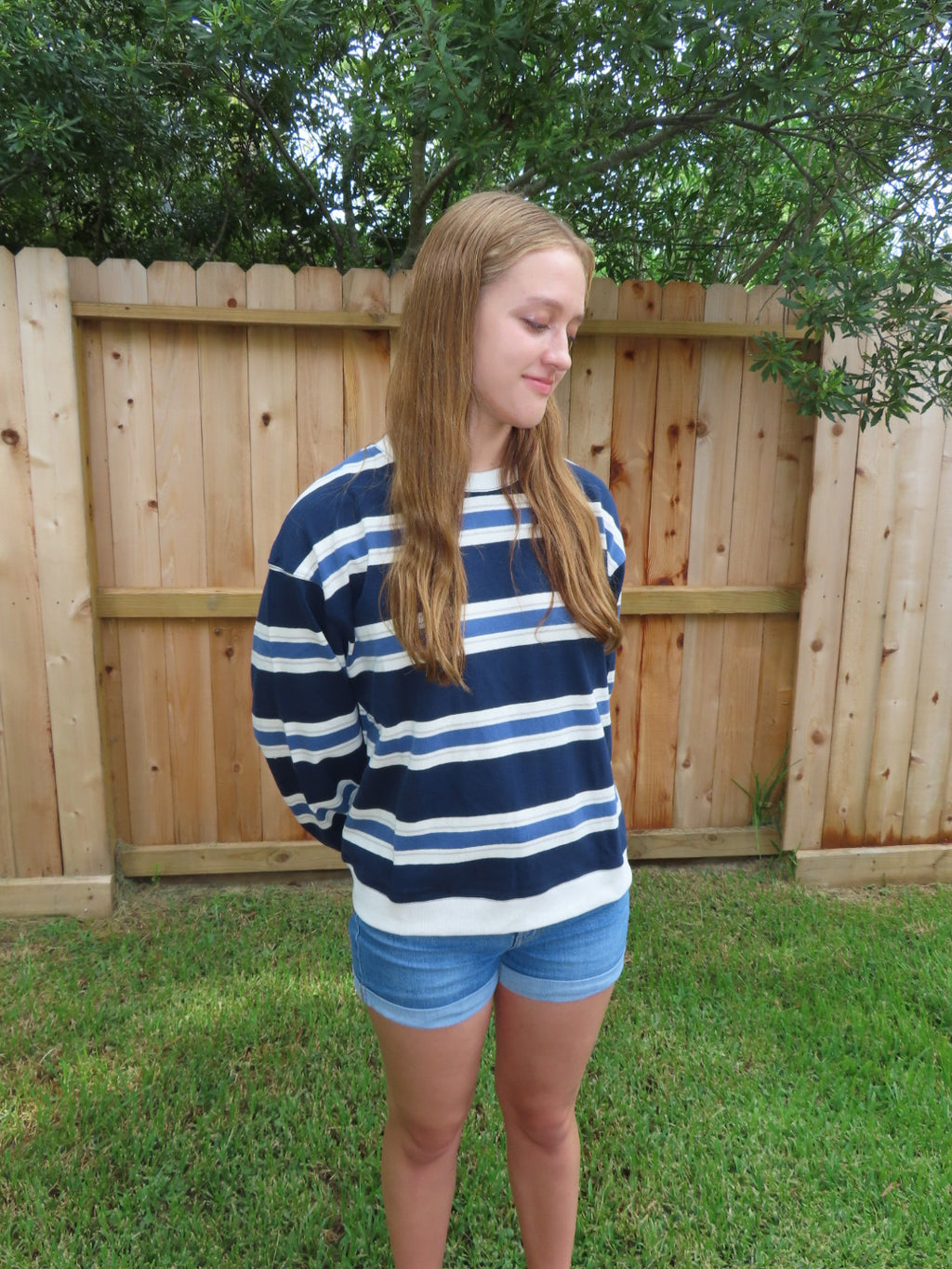 A woman wearing a navy blue and white striped t-shirt and denim shorts, standing outdoors in front of a wooden fence and green grass.