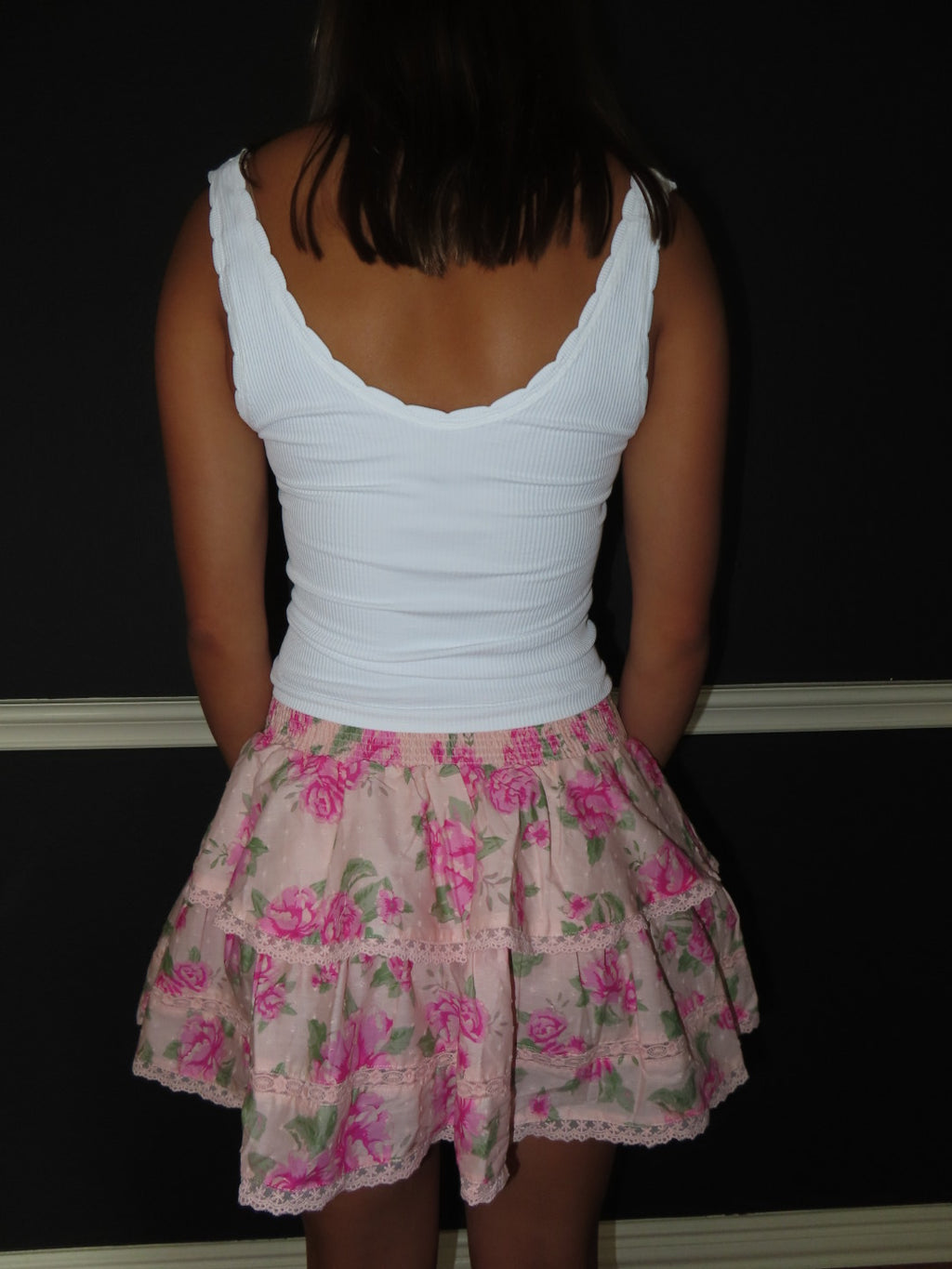 A person shown from the back wearing a white ribbed tank top and a tiered light pink skirt with a pink floral print, standing against a dark wall.