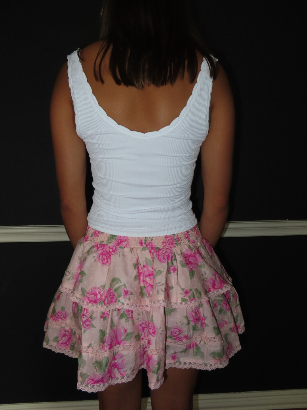 A person shown from the back wearing a white ribbed tank top and a tiered light pink skirt with a pink floral print, standing against a dark wall.