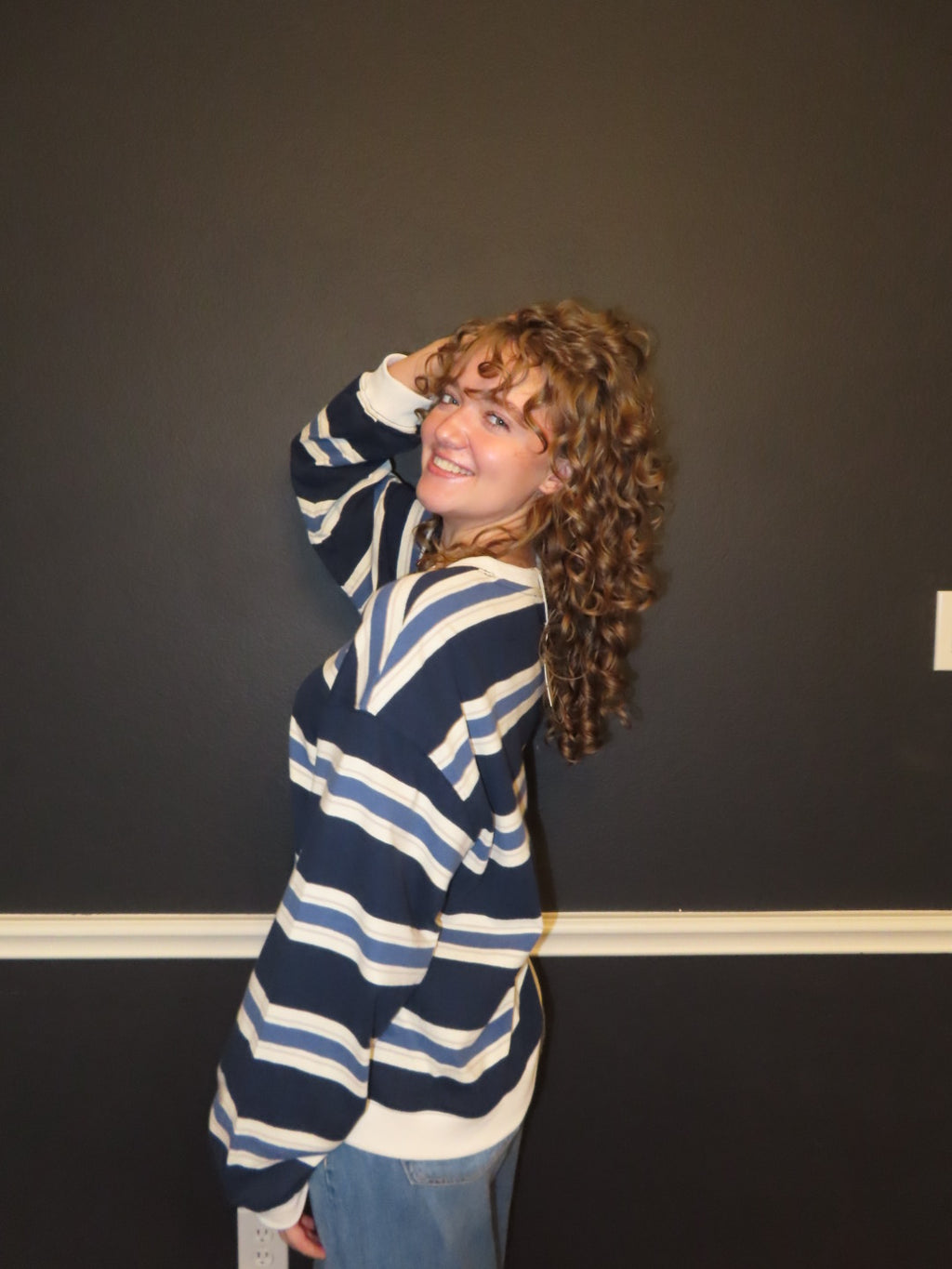A woman with long curly hair wearing an oversized navy and white striped sweater, posing against a dark gray wall.