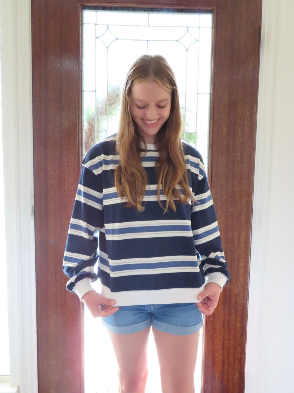A young woman with long light brown hair stands in front of a wooden door wearing a navy blue and white horizontally striped long-sleeve crewneck sweatshirt and denim shorts.