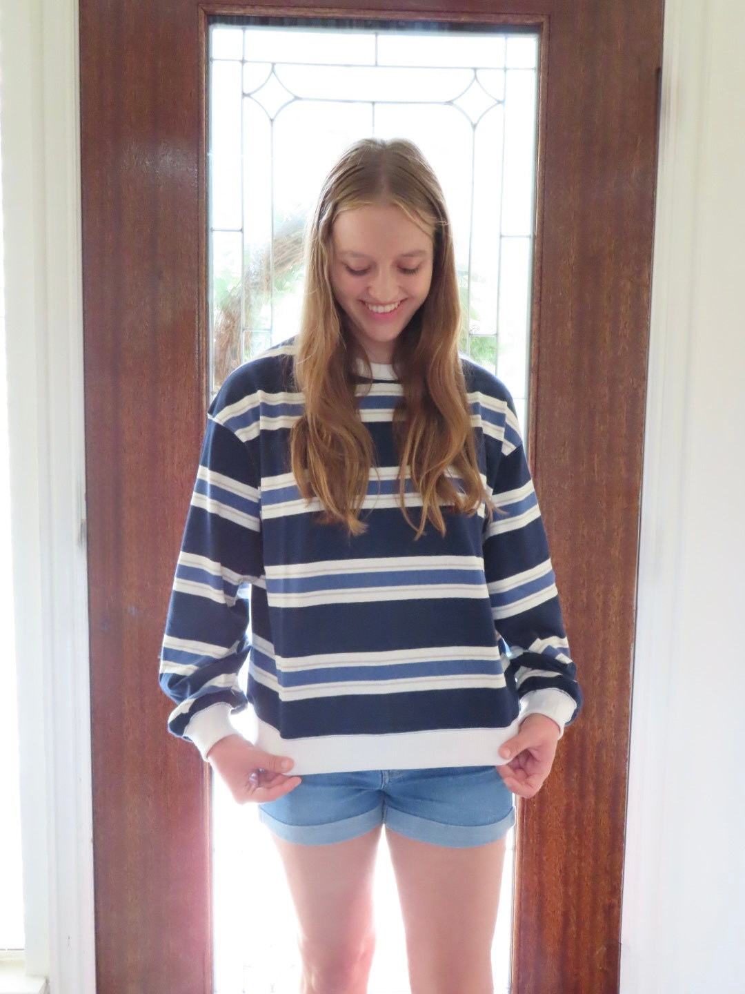 A young woman with long light brown hair stands in front of a wooden door wearing a navy blue and white horizontally striped long-sleeve crewneck sweatshirt and denim shorts.