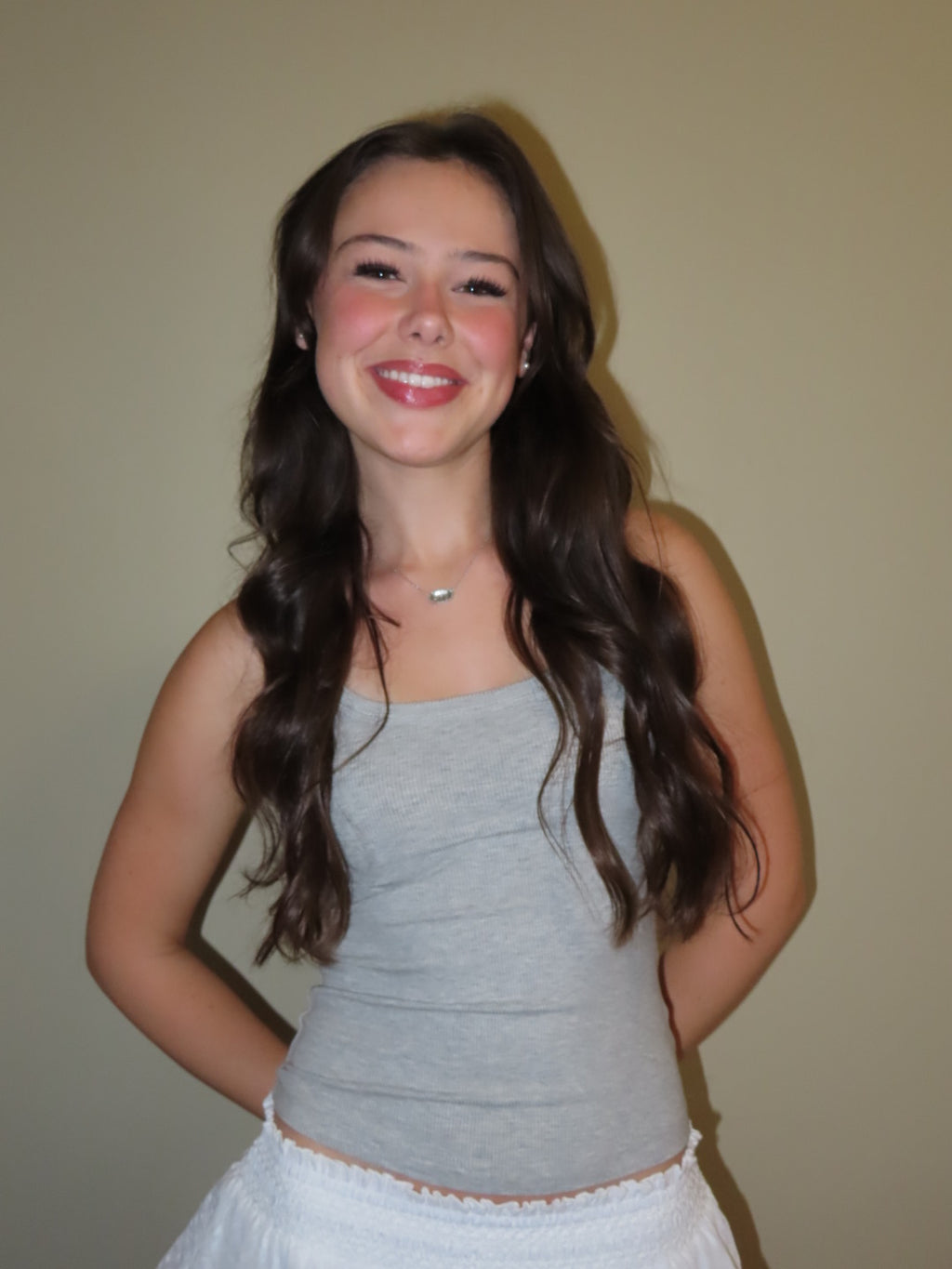 A smiling young woman with long wavy brown hair wearing a light gray ribbed tank top and a white skirt, standing against a plain beige wall.