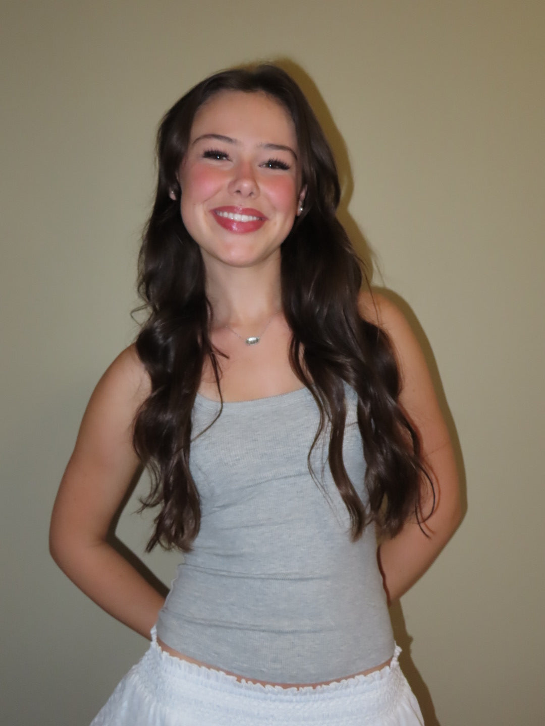 A smiling young woman with long wavy brown hair wearing a light gray ribbed tank top and a white skirt, standing against a plain beige wall.
