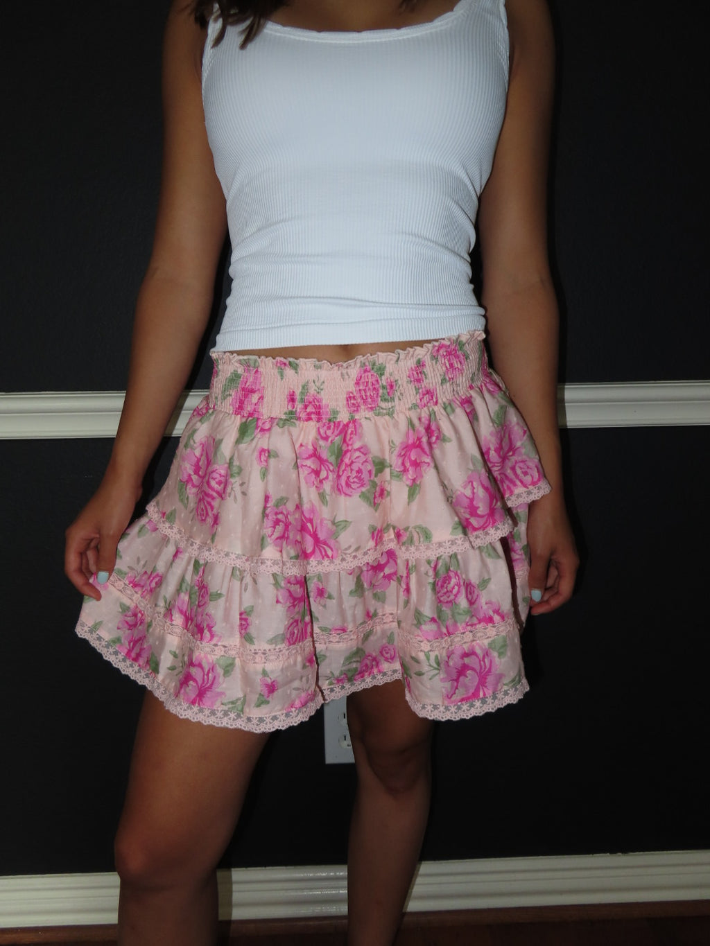A person wearing a white ribbed tank top and a light pink tiered ruffle skirt with a pink rose floral print, standing against a dark wall.