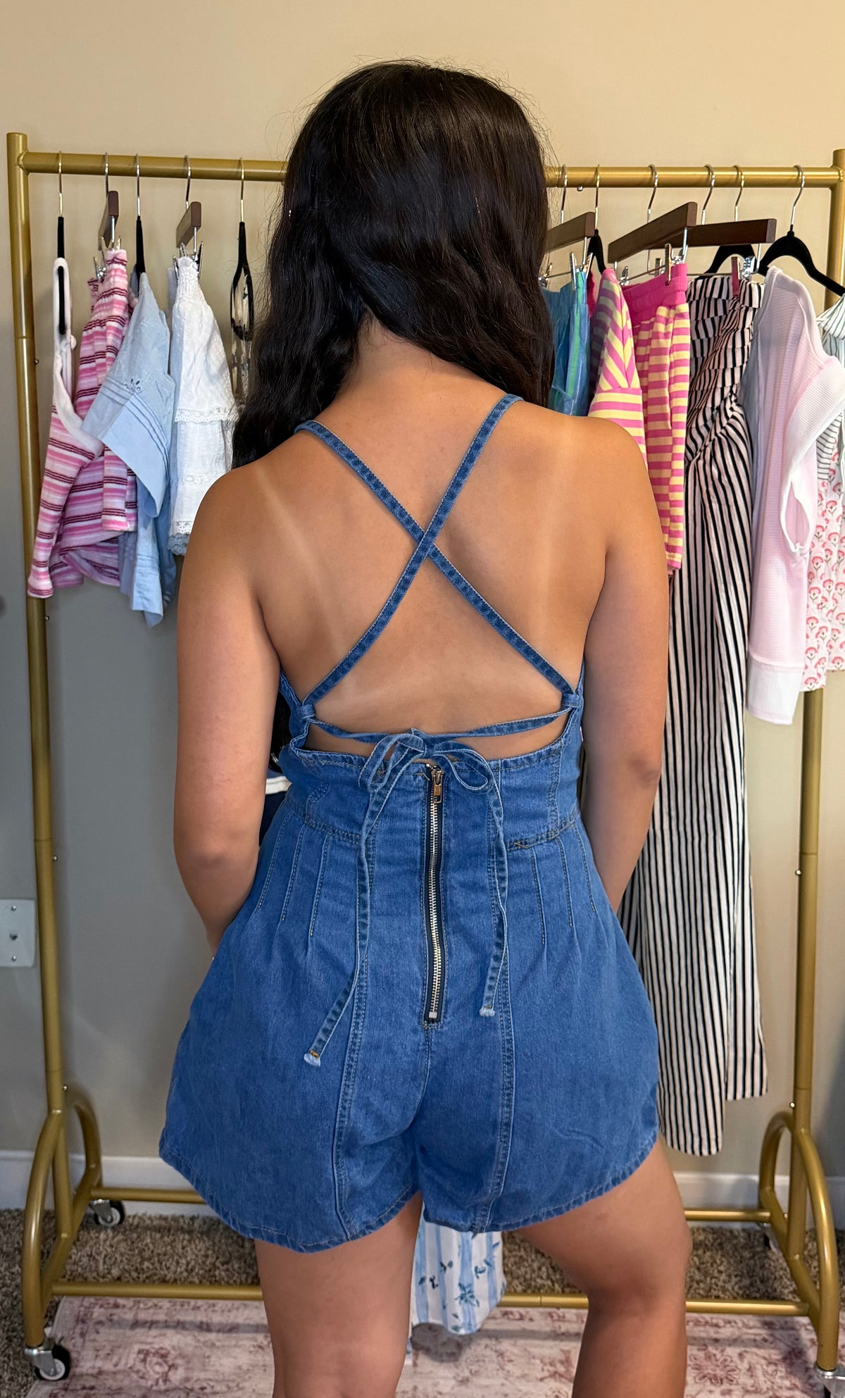 A woman viewed from behind wearing a blue denim romper with a crossover crisscross back strap design and a zippered back. She is standing in front of a gold clothing rack filled with various garments.