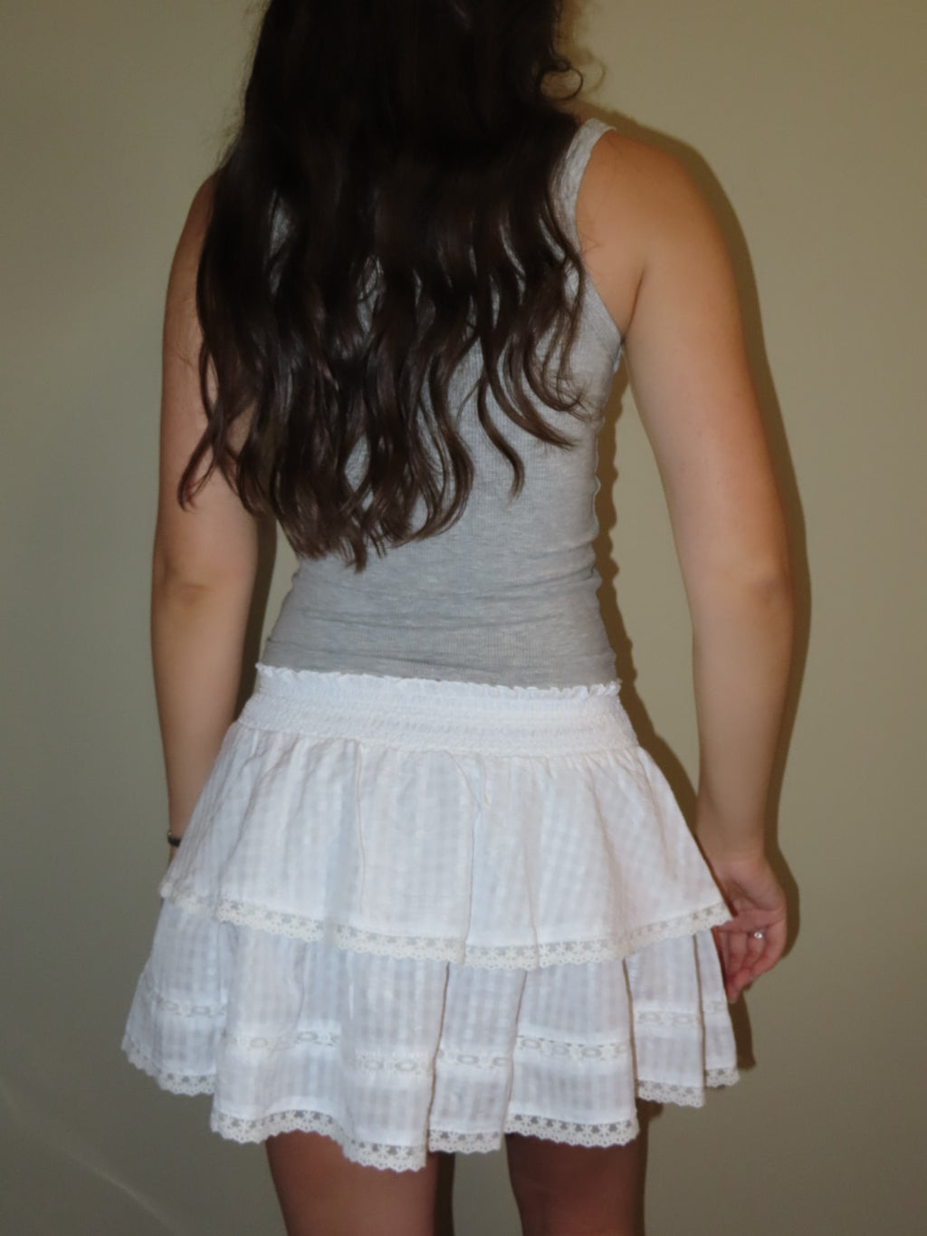 A woman shown from behind wearing a white tiered skirt with eyelet lace trim and a gray tank top, standing against a plain beige wall.