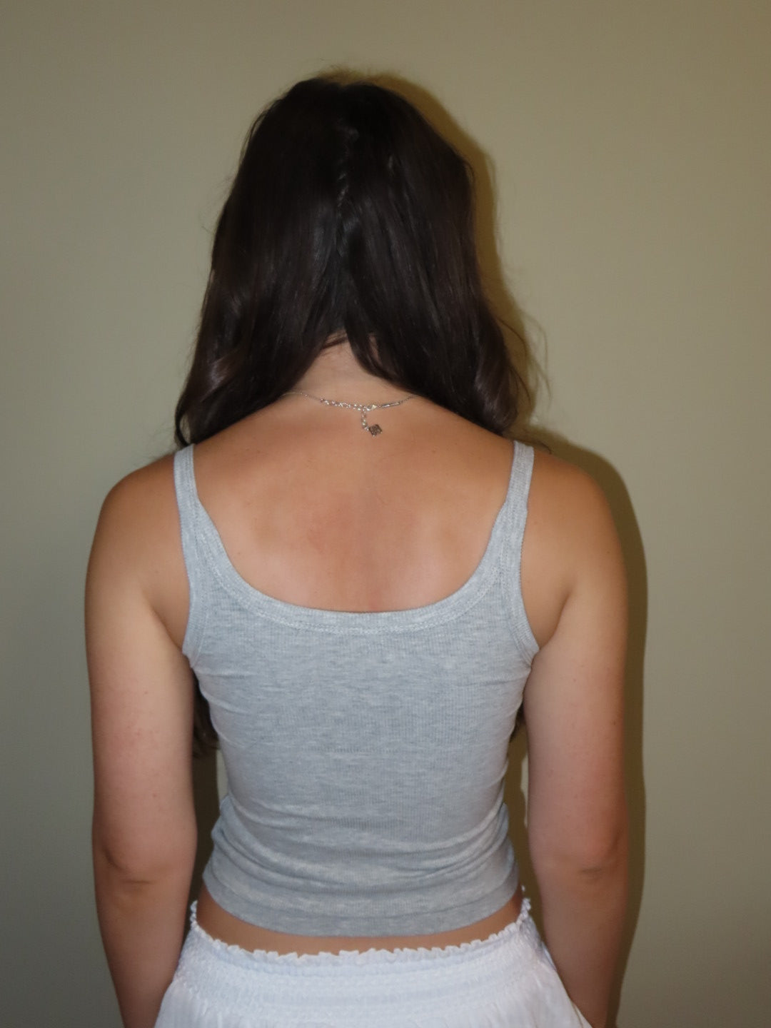 A woman viewed from behind wearing a light gray ribbed tank top and white bottoms, standing against a plain beige wall.