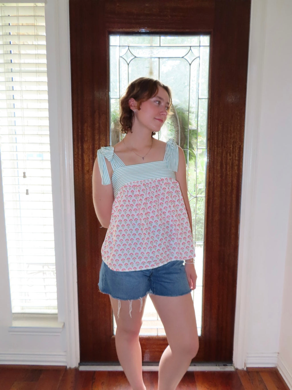 A young woman stands in front of a dark wood door wearing a light green and pink patterned sleeveless top with ruffled straps and blue denim shorts.