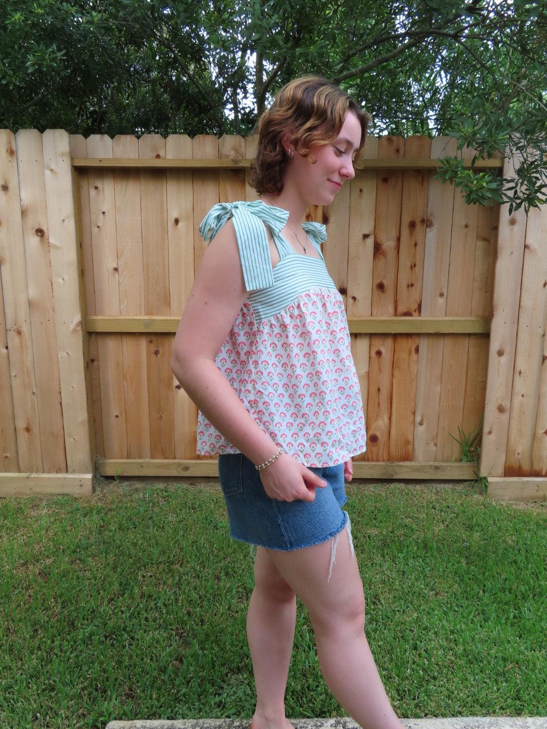 A woman wearing a sleeveless top with a pink and white floral pattern and a light green and white striped straps, paired with denim shorts, standing outdoors in front of a wooden fence.