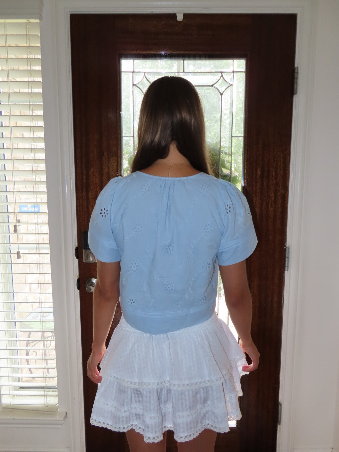 A woman viewed from behind wearing a light blue short-sleeve eyelet top and a white tiered ruffled skirt, standing in front of a dark brown wooden front door.