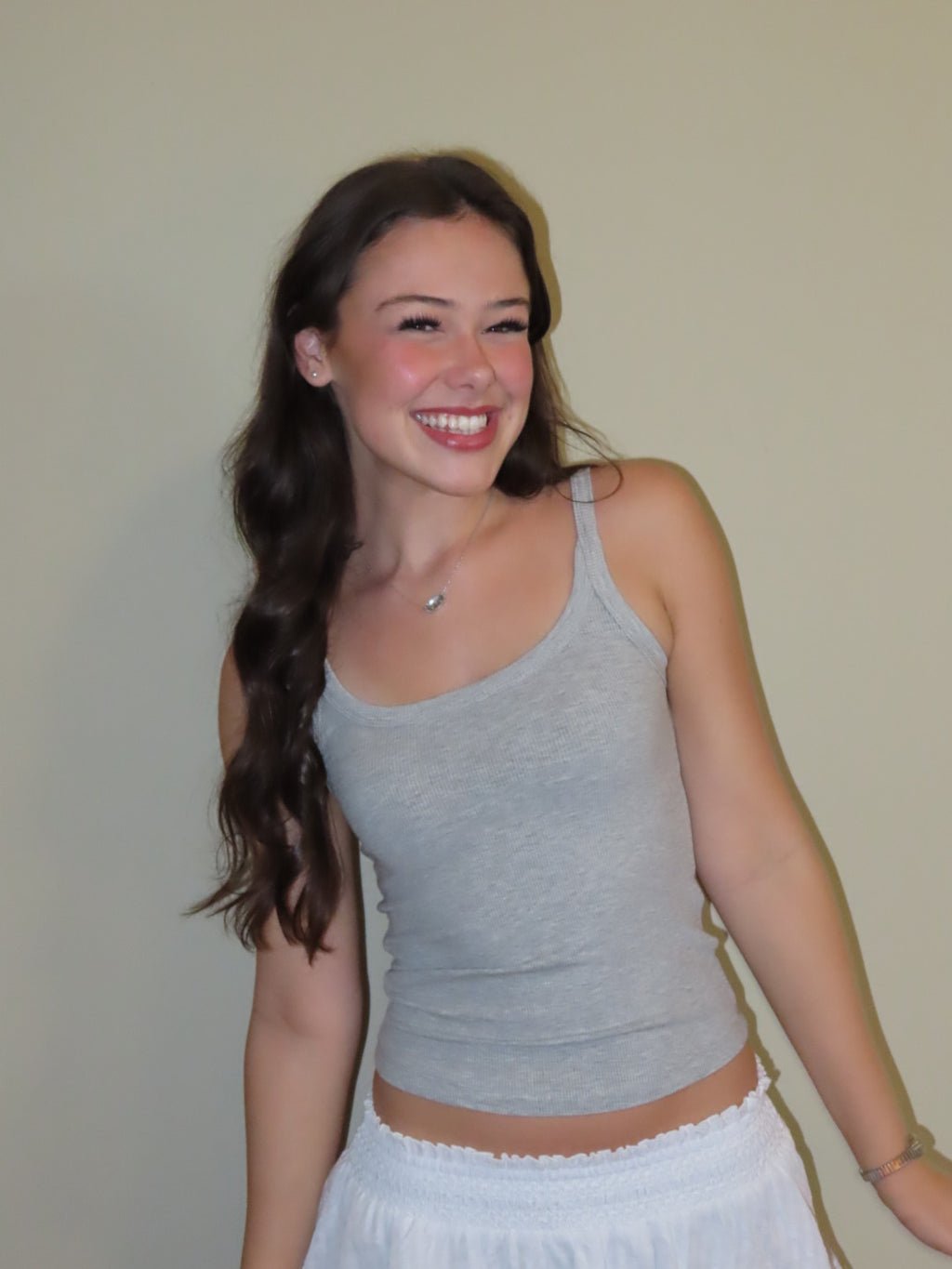 A smiling young woman with long dark hair wearing a light gray ribbed tank top and a white skirt, standing against a plain beige wall.