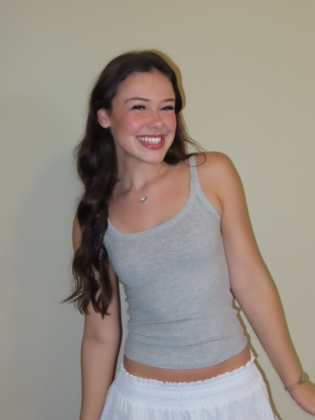 A smiling young woman with long dark hair wearing a light gray ribbed tank top and a white skirt, standing against a plain beige wall.