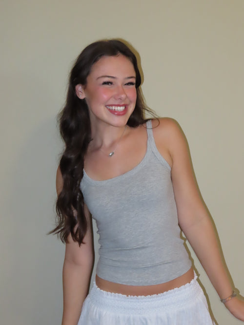 A smiling young woman with long dark hair wearing a light gray ribbed tank top and a white skirt, standing against a plain beige wall.