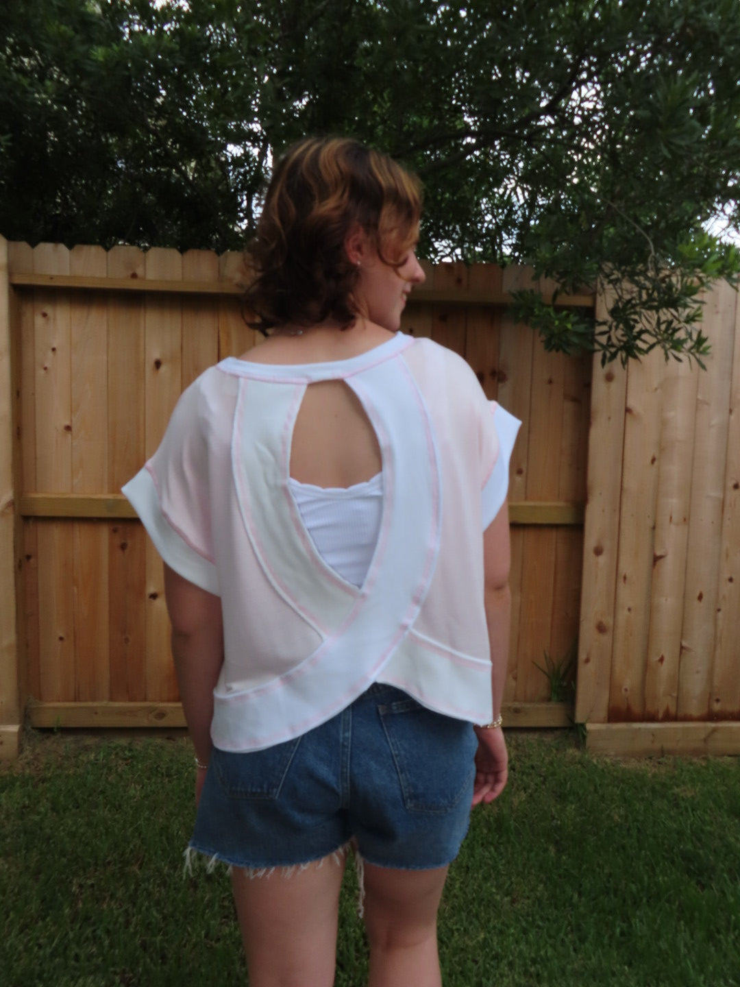 A person wearing a pink short sleeve top with a large open back cutout and denim shorts, standing outdoors in front of a wooden fence and trees.