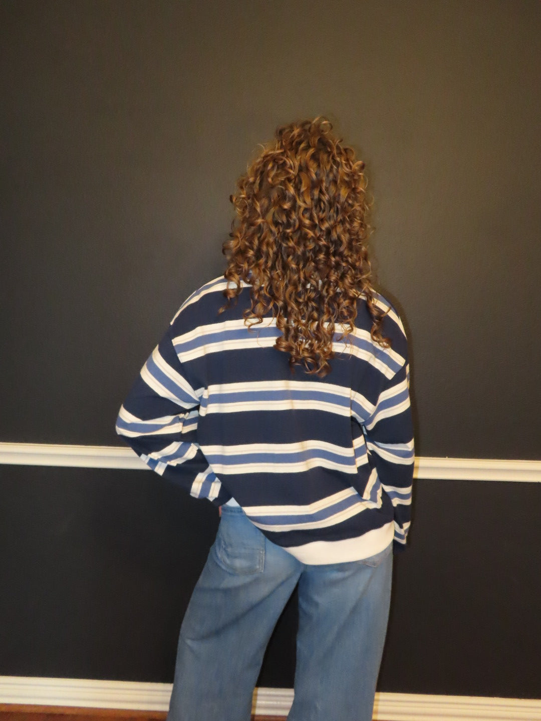 A person viewed from behind wearing a navy blue and white striped long-sleeve shirt and light blue jeans, standing against a dark gray wall with white baseboard molding.