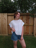 A woman wearing a pink relaxed fit shirt and denim shorts, standing outdoors in front of a wooden fence and green trees.