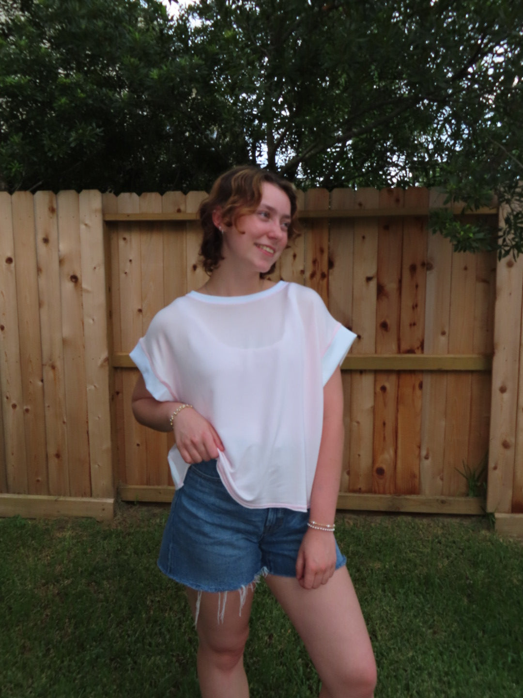 A woman wearing a pink relaxed fit shirt and denim shorts, standing outdoors in front of a wooden fence and green trees.