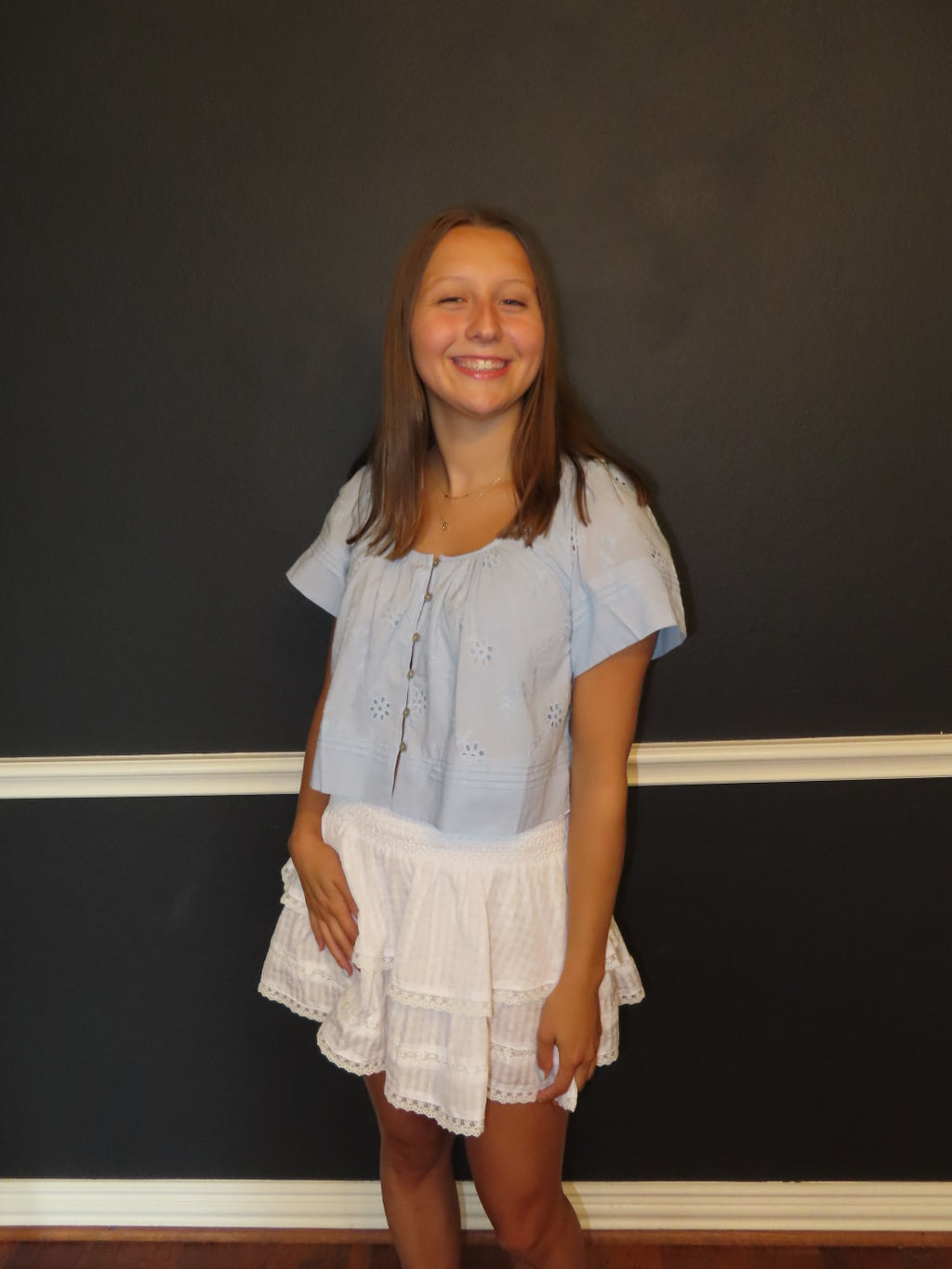A young girl with long light brown hair stands smiling in front of a dark charcoal wall with white trim. She is wearing a light blue eyelet short-sleeve button up blouse and a white tiered ruffled skirt.
