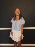 A young girl with long light brown hair stands smiling in front of a dark charcoal wall with white trim. She is wearing a light blue eyelet short-sleeve button up blouse and a white tiered ruffled skirt.