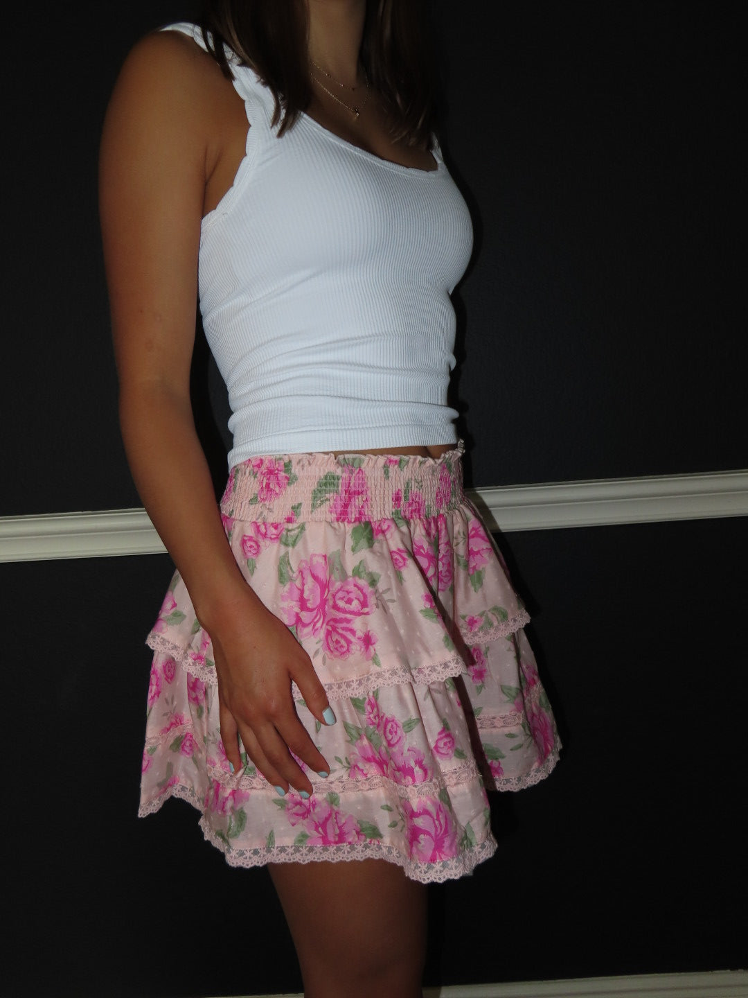 A woman wearing a white ribbed tank top and a light pink tiered skirt with a pink rose floral print and lace trim, standing against a dark background.