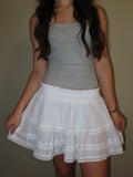 A woman wearing a gray sleeveless tank top and a white tiered ruffle skirt with lace trim, standing against a plain beige wall.