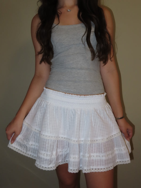 A woman wearing a gray sleeveless tank top and a white tiered ruffle skirt with lace trim, standing against a plain beige wall.